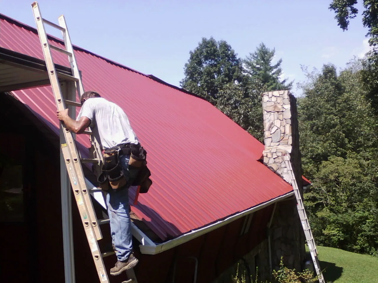 Roofer on a ladder beside a red metal roof and stone chimney — Soffit & Fascia Repair by Roofing Brothers in Startex